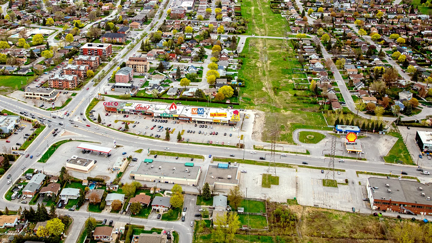 Aerial view of Les Promenades Bellerose, a commercial shopping plaza situated at a busy urban intersection. The plaza features a long, single-story retail strip with visible storefront signs for businesses such as CIBC, Subway, A&W, The Beer Store, M&M Food Market, Bulk Barn, and Dollarama. In front of the plaza is a large surface parking lot with many parked cars. Adjacent to the shopping area is a Shell gas station with a bright yellow and red canopy. Surrounding the plaza are residential neighborhoods with a mix of houses and low-rise apartment buildings, as well as a large green utility corridor running behind the plaza.
