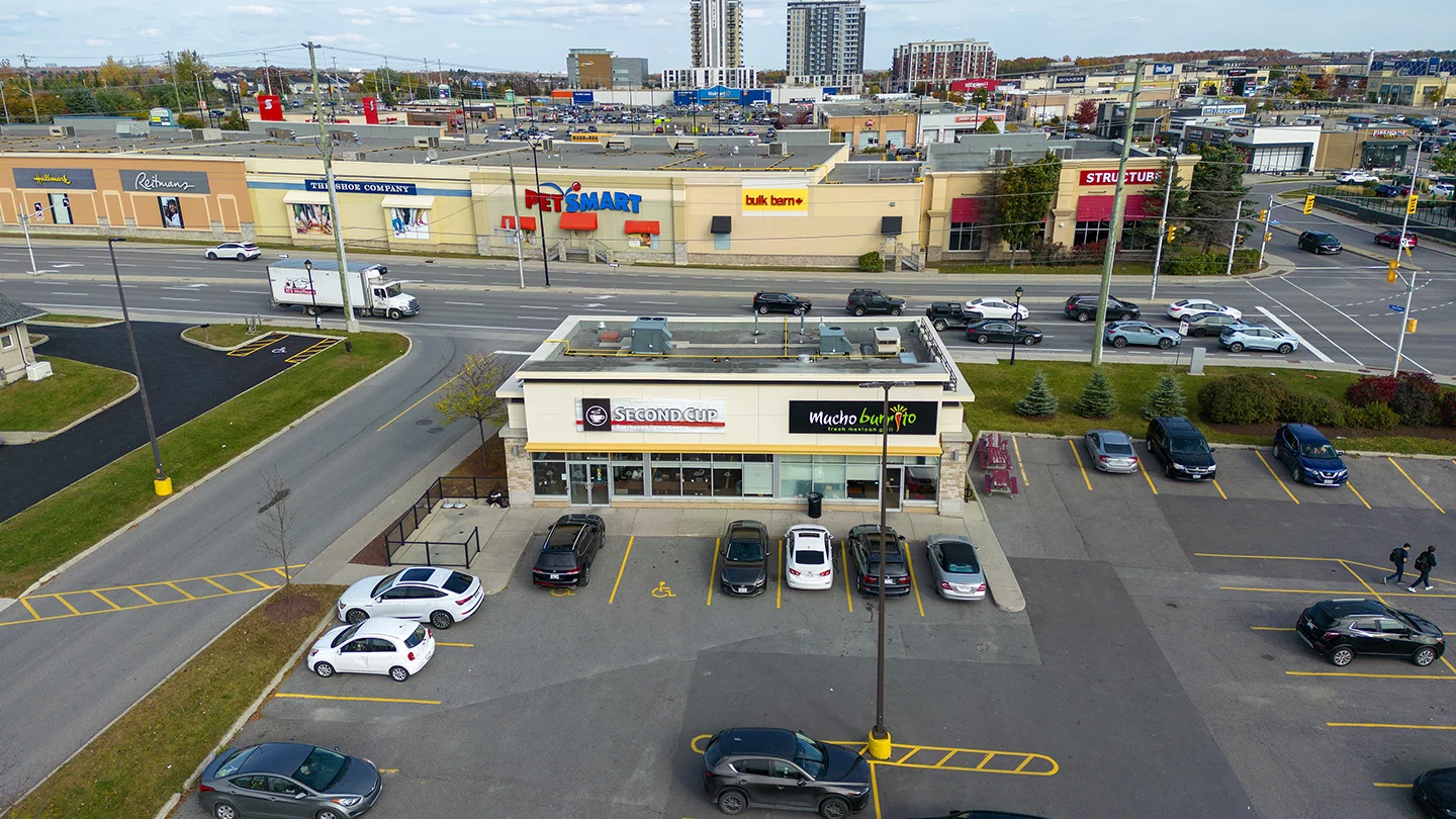 A wide aerial view of Barrhaven Town Centre, showing a standalone retail building in the foreground with signage for Second Cup and Mucho Burrito. Several vehicles are parked in front of the building, and a few pedestrians are walking nearby. Across the street, a larger retail plaza includes stores like PetSmart, Bulk Barn, Reitmans, Hallmark, The Shoe Company, and The Structube furniture store. The scene is framed by multiple roads with moving traffic, traffic lights, and commercial buildings stretching into the background, including mid-rise residential towers under construction.
