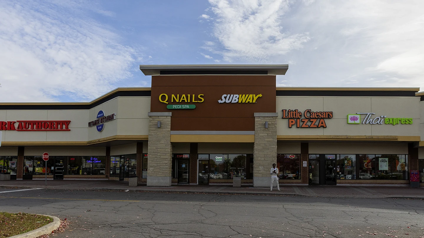 A street-facing view of a retail strip plaza in Barrhaven Town Centre featuring a variety of storefronts. From left to right, visible businesses include Q Nails Pedi Spa, Subway, Little Caesars Pizza, and Thai Express. A person in white clothing stands on the sidewalk in front of the stores. The building has a neutral beige and brown color scheme with stone pillars and large glass windows. A cracked asphalt parking area and curb with some fallen leaves are in the foreground, under a partly cloudy sky.