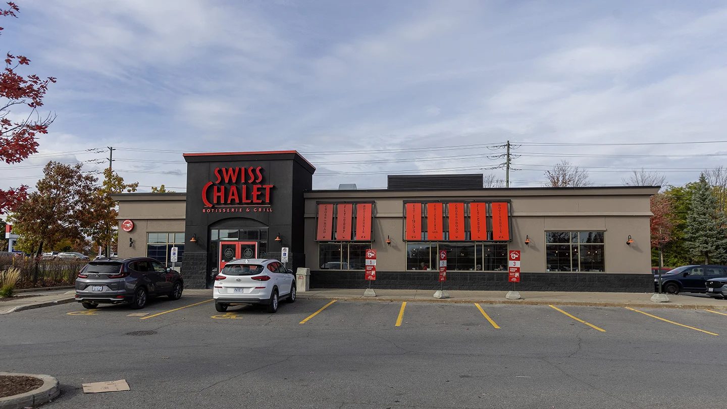 Exterior view of a Swiss Chalet Rotisserie & Grill restaurant in Barrhaven Town Centre. The building has a modern design with dark and neutral tones, featuring bold red signage and matching red awnings above the windows that display menu highlights. The restaurant entrance is centered under a black facade with two accessible parking spaces in front. Several cars are parked in the lot, and red maple trees with autumn leaves frame the scene on the left. The sky is mostly cloudy with a few patches of blue.