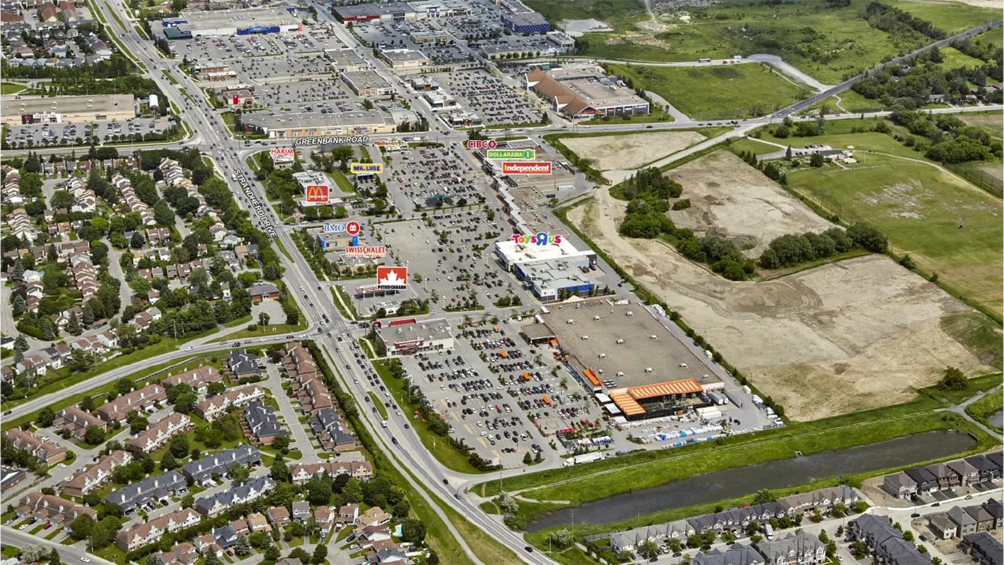 A detailed aerial image showing a large suburban commercial and residential area in Barrhaven. In the center, a major intersection connects Greenbank Road and Strandherd Drive, surrounded by various retail stores, including prominent brands with large, visible signage: Home Depot, Toys “R” Us, Independent, Dollarama, Petro-Canada, Swiss Chalet, McDonald’s, BMO Bank, Mr. Lube, and CIBC. Parking lots are full of vehicles, and strip plazas extend outward in multiple directions. On the left, residential neighborhoods with tree-lined streets and single-family homes are visible. To the right and background, large open fields and green spaces border the developed area, indicating future potential expansion or preserved land.