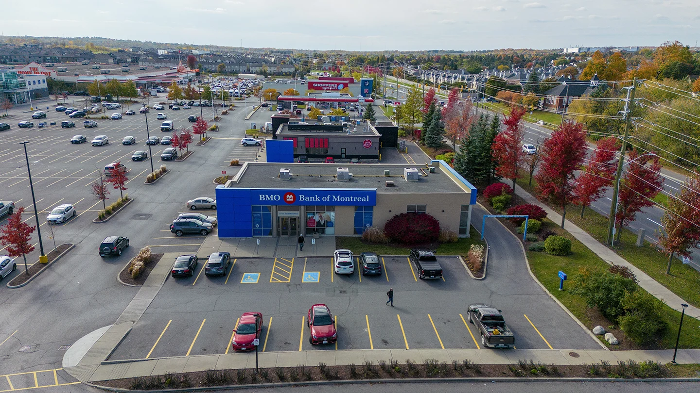 Aerial view of a BMO Bank of Montreal branch located in a commercial plaza in Barrhaven Town Centre. The bank building features a blue exterior with the BMO logo above the main entrance. Several vehicles are parked in front, including accessible parking spaces marked in yellow. Behind the bank is a Swiss Chalet restaurant, and in the distance, a Shoppers Drug Mart can be seen along with a large Home Depot. To the right, a tree-lined street is filled with vibrant red and orange autumn foliage, and residential houses are visible beyond. Power lines run parallel to the sidewalk on the right side of the image.
