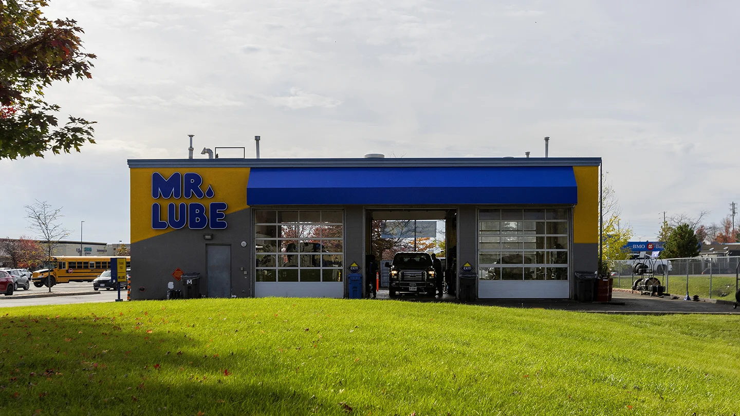 Front view of a Mr. Lube automotive service centre in Barrhaven Town Centre, with a bold yellow and blue exterior. The building has three large glass garage doors, one of which is open with a black pickup truck partially inside, receiving service. "MR. LUBE" is written in large, dark blue letters on the top-left corner of the building. A grassy area is in the foreground, and in the background, a school bus, parked vehicles, and signage for a BMO Bank of Montreal branch are visible under a bright, cloudy sky.
