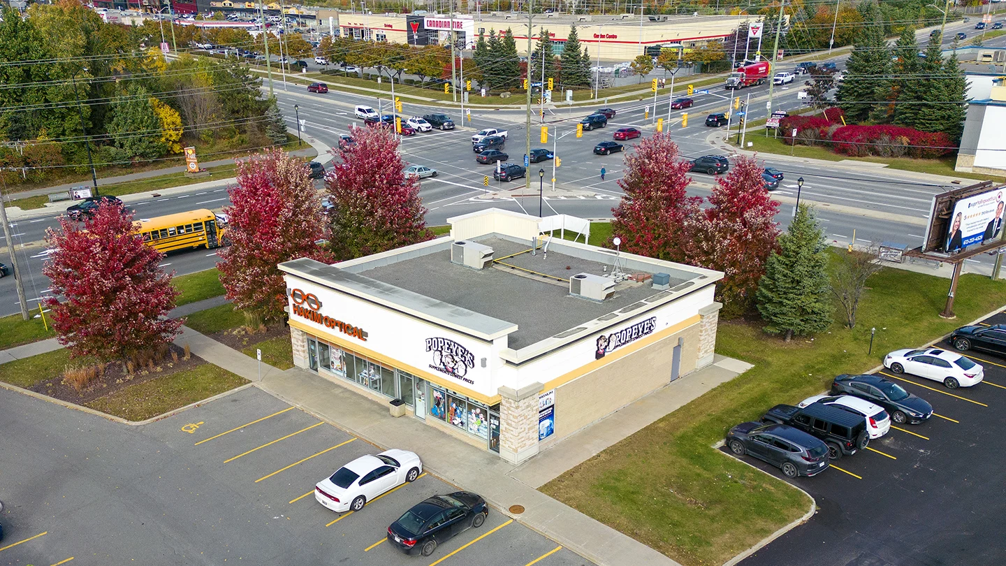 Aerial view of a commercial corner plaza in Barrhaven Town Centre featuring Popeyes Louisiana Kitchen and Hakim Optical. The single-story building has a beige brick and white façade with clear store signage. Surrounding the building is a parking lot with cars and accessible parking spaces, bordered by a sidewalk and red-leafed trees. In the background, a busy intersection is visible with cars, a yellow school bus, and a Canadian Tire store across the street. A billboard and landscaped greenery complete the suburban commercial setting under a cloudy sky.