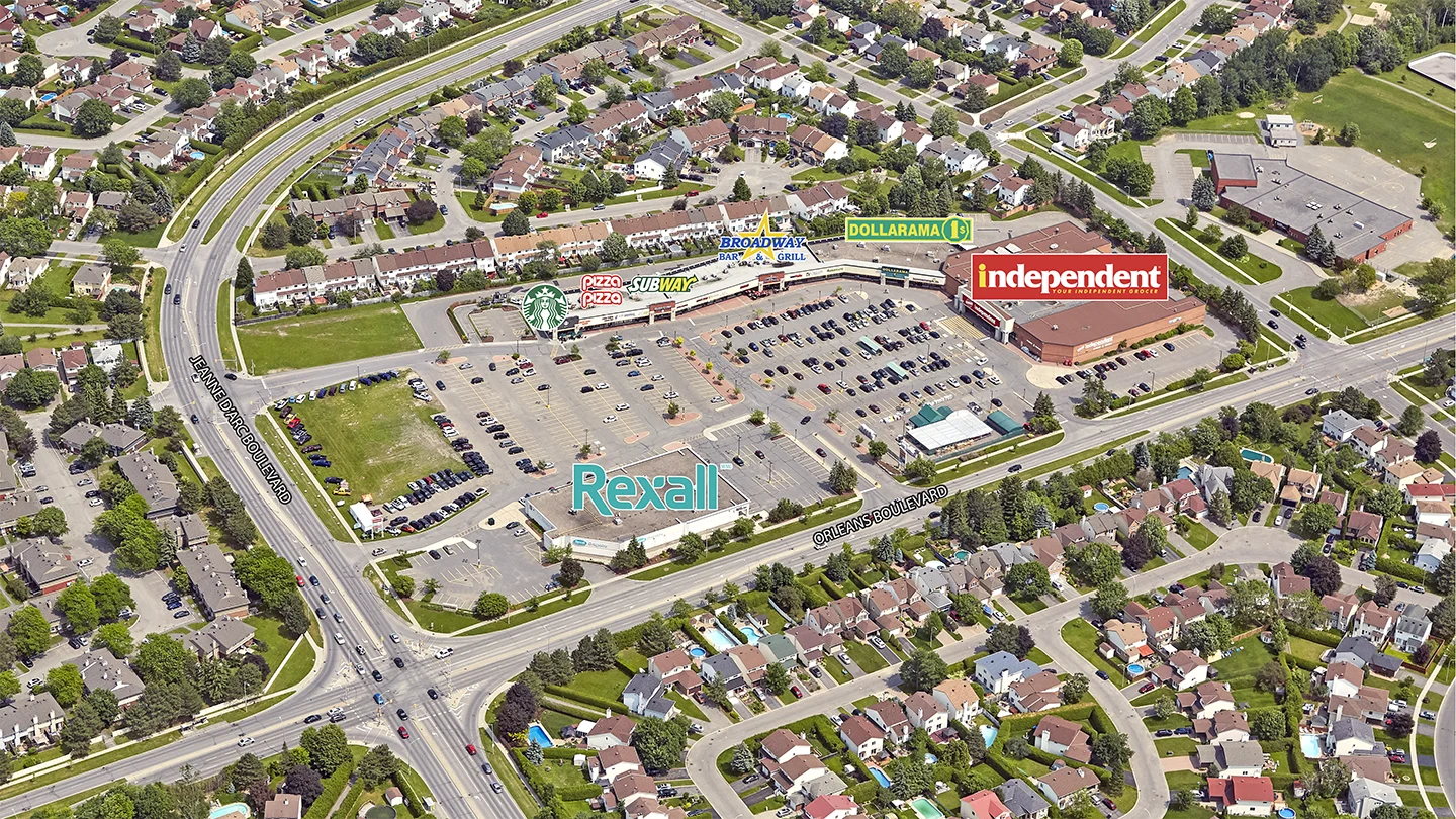Aerial image of the Orleans Gardens Shopping Plaza in Ottawa, Ontario, surrounded by residential homes and intersecting roadways. The plaza features large visible signs for businesses including Independent (a supermarket), Rexall, Dollarama, Subway, Pizza Pizza, Starbucks, and Broadway Bar & Grill. The shopping center is bordered by Jeanne d’Arc Boulevard and Orleans Boulevard, both labeled in the image. The scene includes a large parking lot partially filled with cars and green spaces interspersed among tightly packed suburban homes, many with backyard swimming pools.