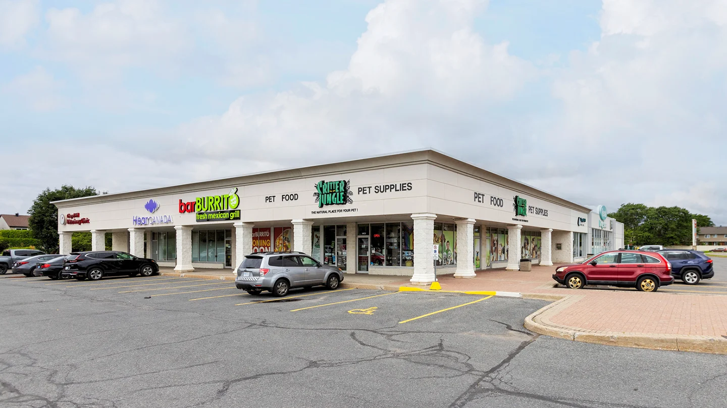 Street-level view of a single-storey strip mall at Orleans Gardens Plaza in Ottawa, Ontario, with clear storefront signs for Critter Jungle (a pet food and supplies store), HearCANADA, barBURRITO (a fresh Mexican grill), and Royal Lepage Performance Realty. The storefronts are framed by white pillars and overhangs, with a visible accessible parking space in front. Several cars are parked in the lot, and the sky is partly cloudy.