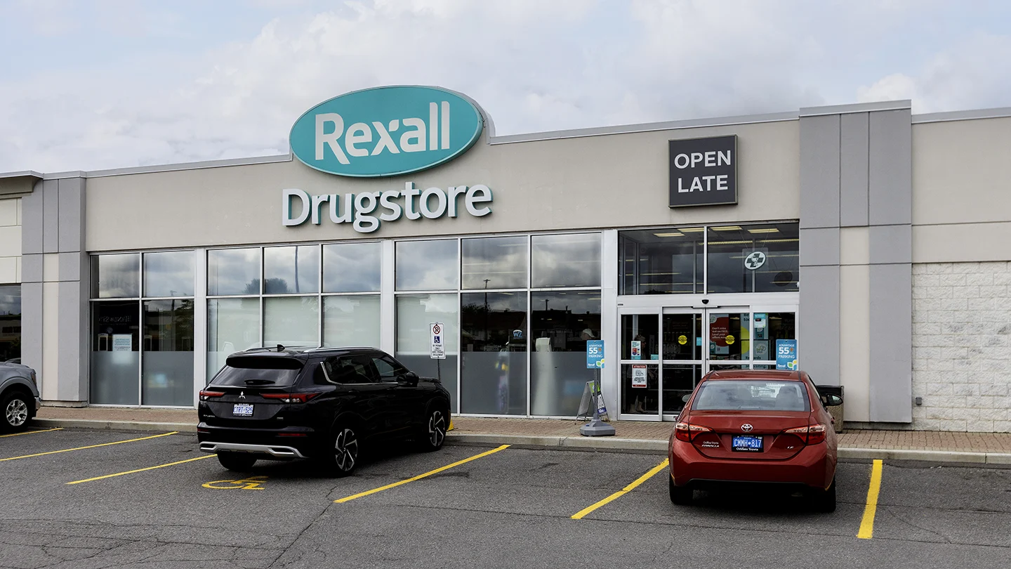 Exterior view of a Rexall Drugstore at Orleans Gardens Plaza in Ottawa, Ontario. The store features a large teal and white Rexall sign above the main entrance, with a smaller grey “Open Late” sign beside it. Two cars are parked in front of the entrance, including one in an accessible parking space. The automatic doors and visible pharmacy signage indicate accessibility and extended hours. The sky above is partly cloudy.