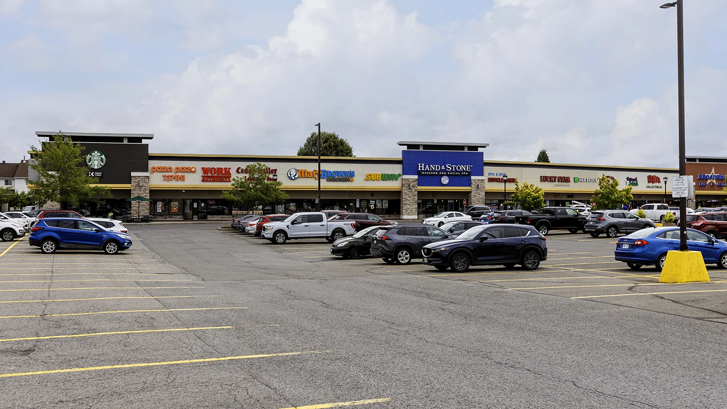 A wide-angle view of a storefront strip at Orleans Gardens Plaza in Ottawa, Ontario, showing a variety of businesses. Visible stores include Starbucks, Pizza Pizza, Work Authority, Cedar Valley Lebanese Food, Mary Brown’s Chicken, Subway, Hand & Stone Massage and Facial Spa, Dollarama, Lucky Star Chinese Food, Kids Klozet, Master Collectibles, and more. The foreground features a large parking lot with numerous parked cars and clear yellow-lined accessible parking spaces. Trees and lampposts are interspersed along the storefronts under a partly cloudy sky.