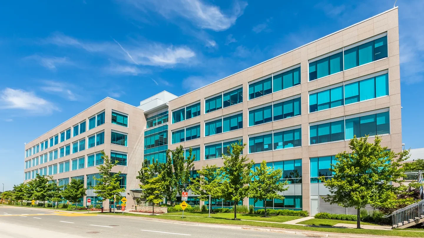 Wide-angle view of a five-storey office building with blue-tinted windows and landscaped grounds, seen on a sunny day with a few clouds in the sky.