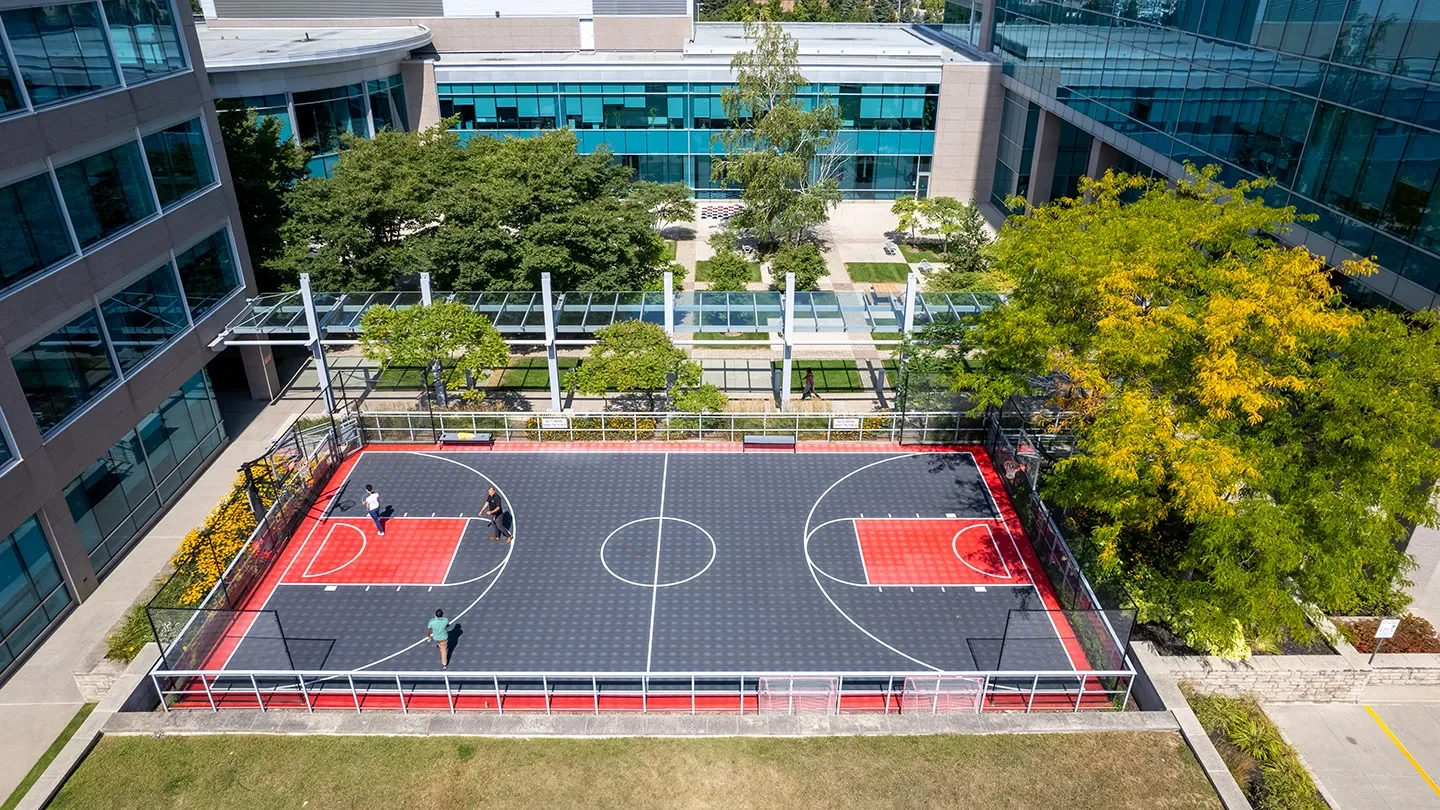 Enclosed outdoor basketball court with four people playing, situated between two glass office buildings with surrounding greenery and walking paths.