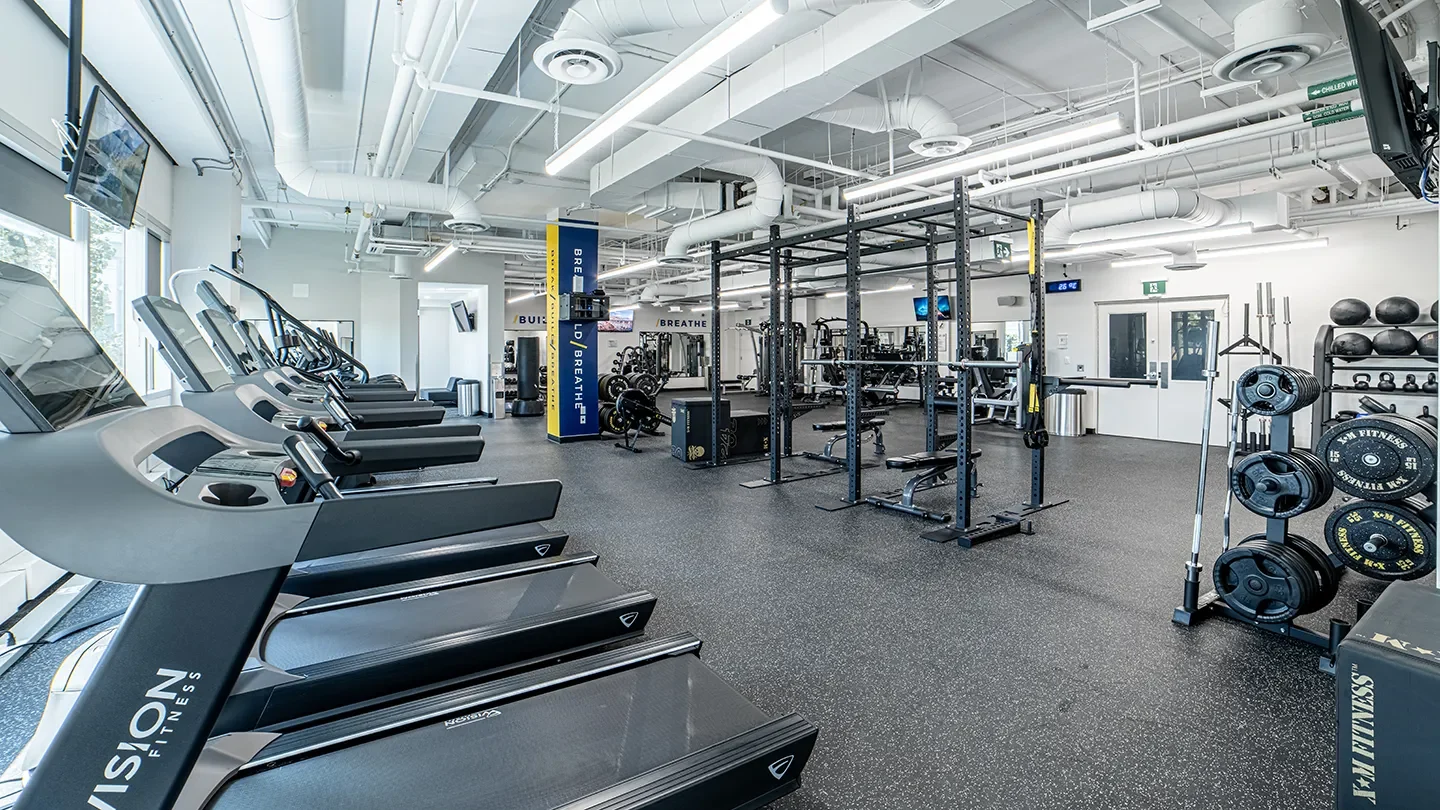 Interior of a well-equipped fitness center featuring treadmills, squat racks, free weights, and strength machines in a brightly lit, modern space with high ceilings.