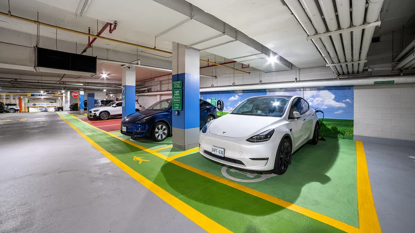 Underground parking garage with brightly lit electric vehicle charging stations, including a white Tesla plugged in and clear green-painted spaces marked for EV use.