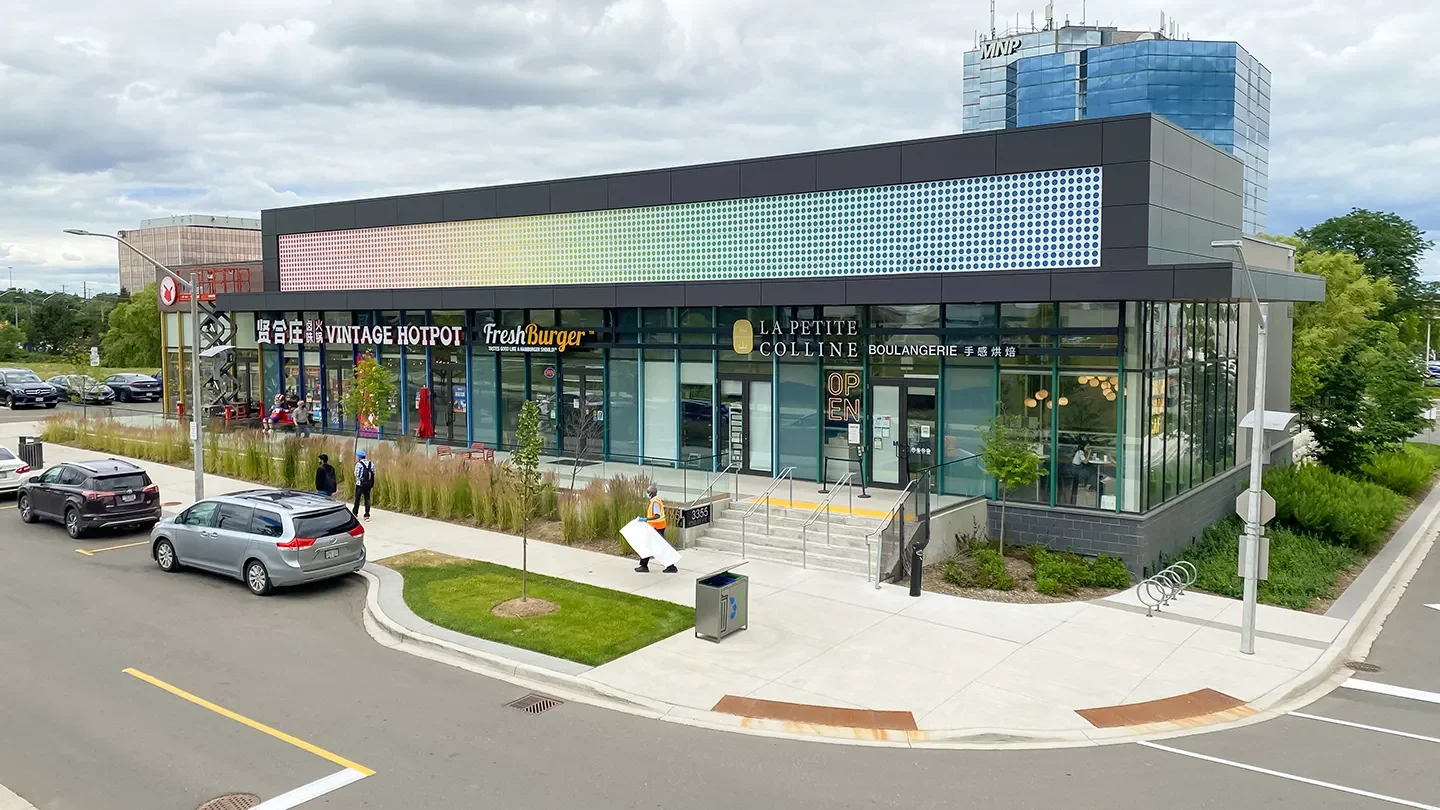 Street-level view of a modern retail plaza featuring businesses like Vintage Hotpot, FreshBurger, and La Petite Colline Boulangerie, with a few pedestrians and cars parked in front.