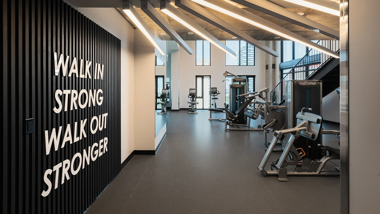 Entrance to a fitness centre with bold white lettering on a black slatted wall reading “WALK IN STRONG, WALK OUT STRONGER,” and a modern gym space visible with exercise machines, tall windows, and angled ceiling lights.