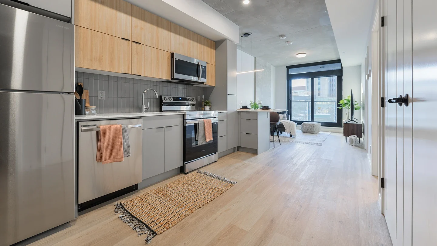 Modern studio apartment interior featuring a sleek kitchen with stainless steel appliances, light wood and grey cabinetry, and a tiled backsplash. The open-concept layout extends to a dining area and living space with large windows and natural light, minimalist decor, and light wood flooring.