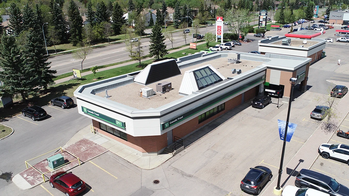 Aerial view of a TD Canada Trust bank branch in a commercial plaza. The building has a flat roof with skylights and HVAC units, green TD signage on the exterior, and a drive-through banking area. Surrounding the bank are multiple parked vehicles, nearby retail businesses, and tree-lined streets.
