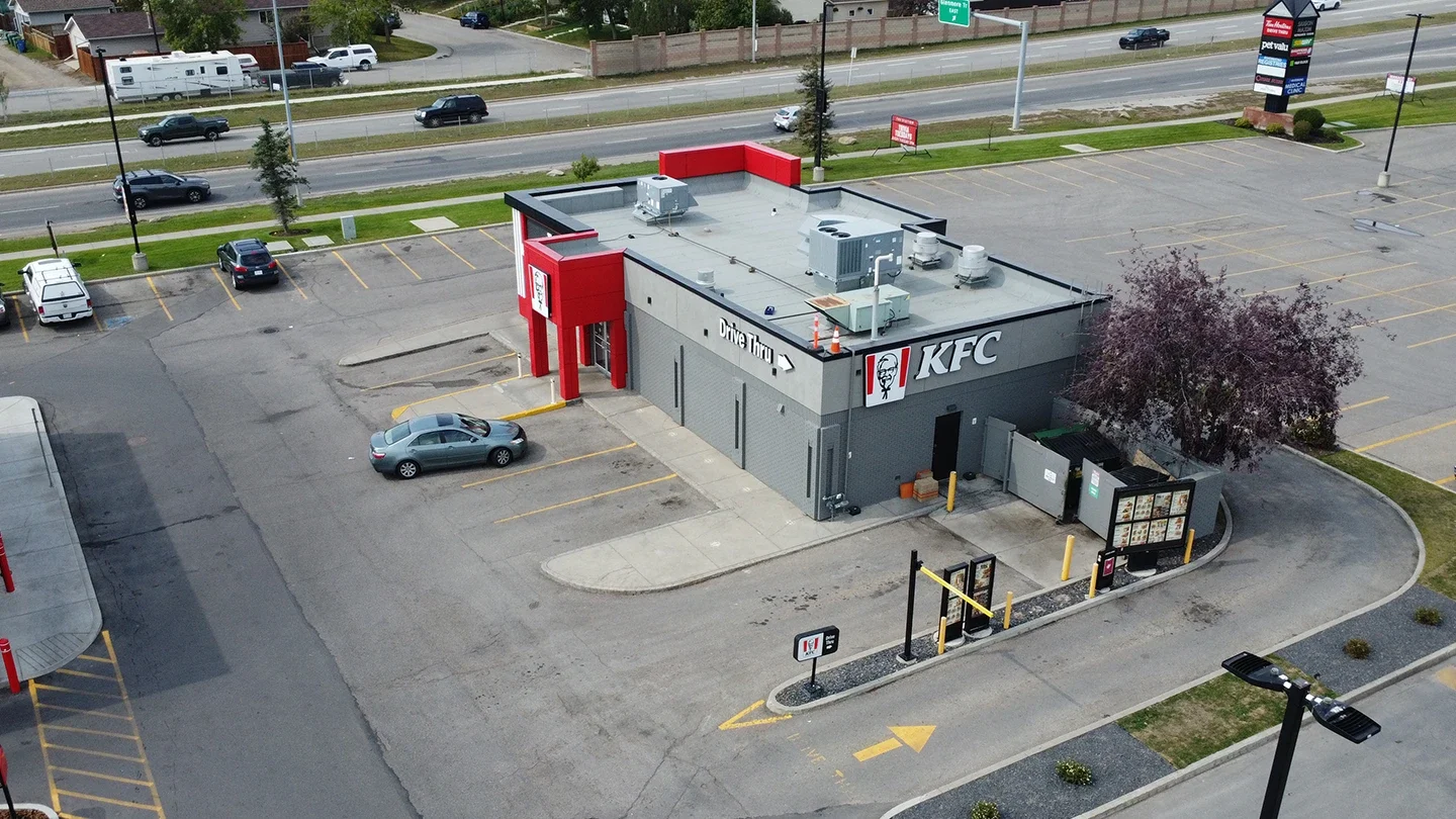 Aerial view of a KFC restaurant with labeled drive-thru lanes, parking spaces, and an accessible sidewalk, situated near a main road with cars and residential homes in the background.
