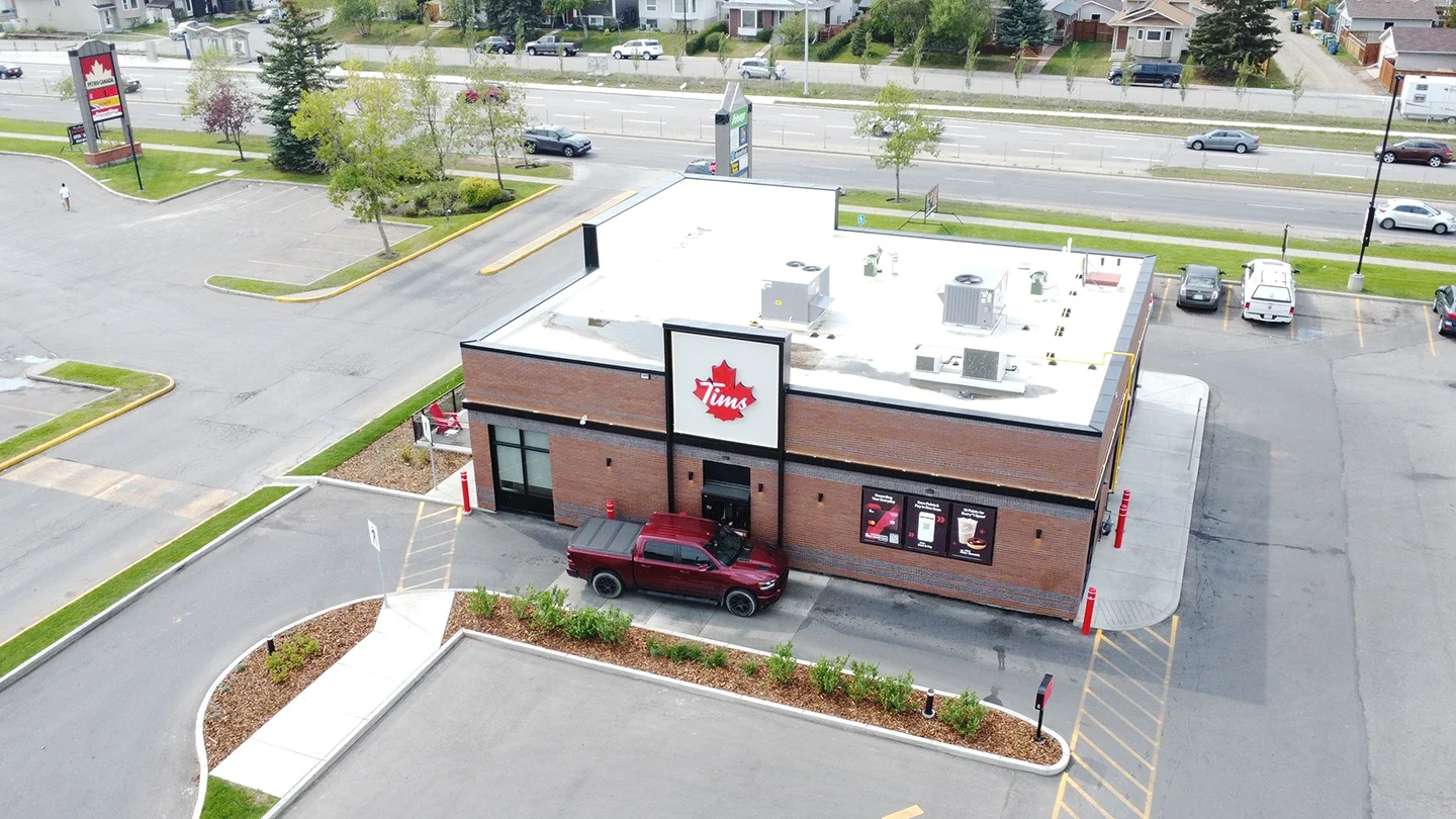 Aerial view of a Tim Hortons building with a red pickup truck parked outside, visible drive-thru lanes, and accessible sidewalk ramps, surrounded by a parking lot and nearby street.