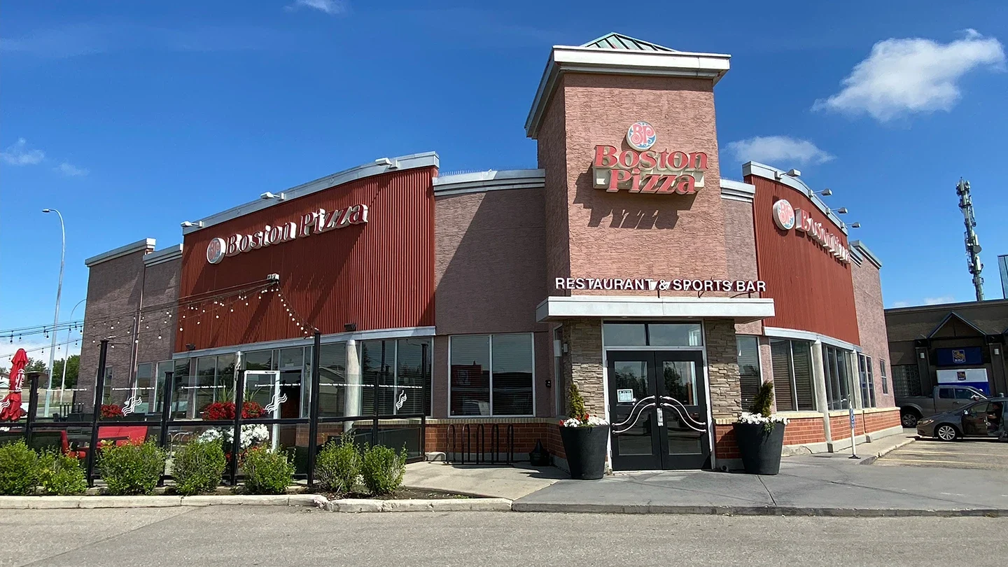 Exterior of Boston Pizza at Riverbend Shopping Centre, featuring red paneling, large windows, a patio with red umbrellas, and the restaurant