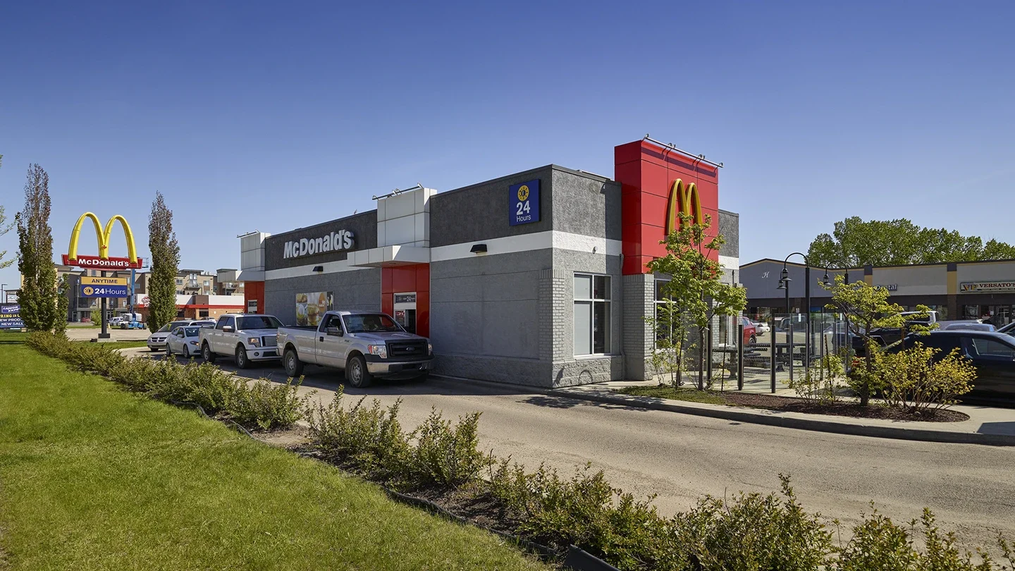 Ground-level view of a McDonald’s restaurant with a drive-thru, featuring a line of vehicles waiting. The building has a grey and red exterior with a 24-hour sign. Other retail storefronts and trees are visible in the background under a clear blue sky.
