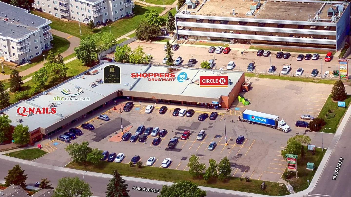 Aerial view of Belmead Shopping Centre, a single-story retail plaza with a flat roof and a large parking lot. A Pepsi truck is parked at the loading area, and several cars are in marked parking spaces. Surrounding the plaza are trees, residential homes, and an office building in the background.