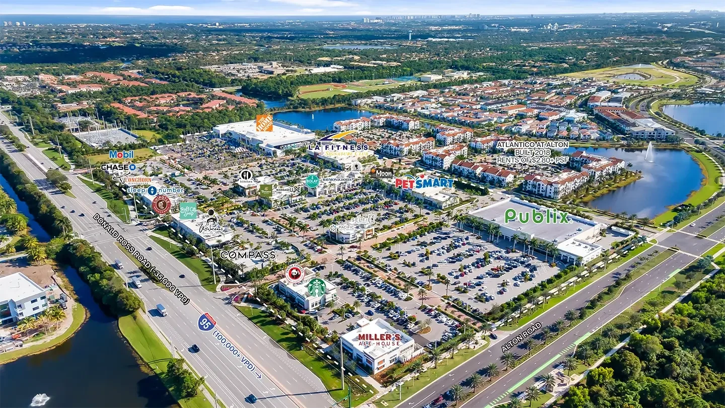 Aerial view of Alton Town Center in Palm Beach Gardens, Florida, showcasing a large mixed-use commercial development. The image highlights labeled retail and service tenants including Publix, LA Fitness, PetSmart, The Home Depot, Starbucks, Panda Express, and Miller’s Ale House. Residential buildings with red-tiled roofs and landscaped lakes surround the shopping area. Major roads such as I-95 and Donald Ross Road are visible with labeled traffic volumes, and the scene includes parking lots, palm-lined streets, and green space throughout.