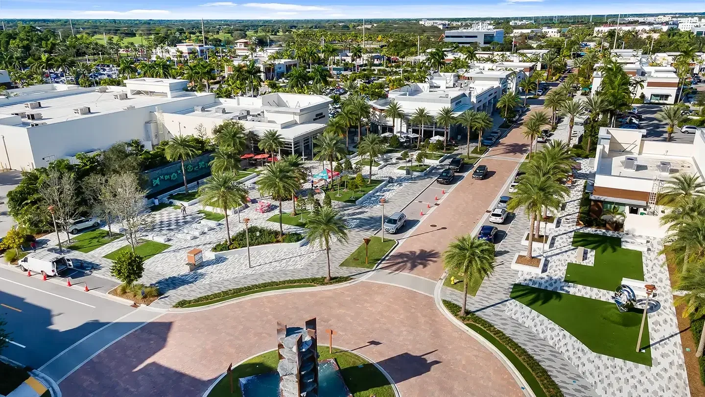 Vibrant pedestrian plaza at Alton Town Center featuring palm trees, patterned walkways, and landscaped green spaces. Modern buildings with outdoor seating and umbrellas line the street, while cars are parked along the road. A roundabout with a contemporary fountain sculpture is visible in the foreground.
