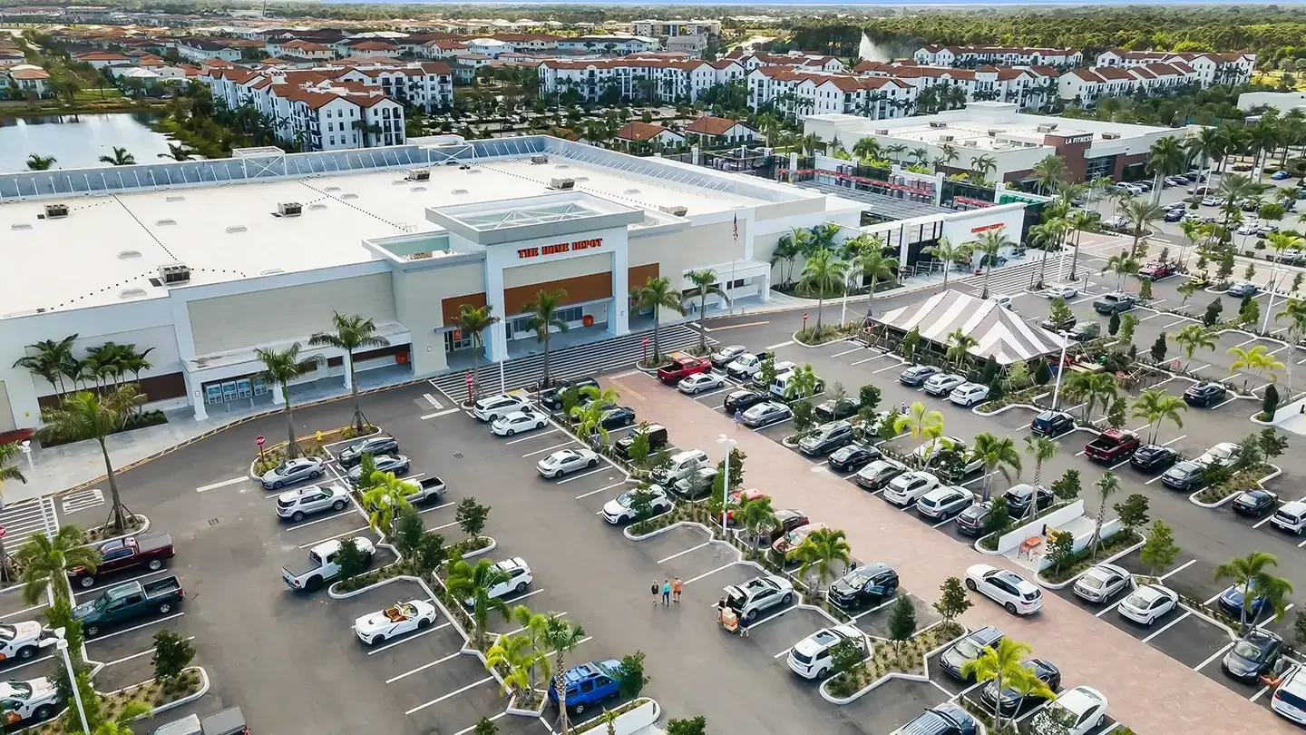 Elevated view of The Home Depot at Alton Town Center, with a large parking lot filled with cars in the foreground. The store has a white and beige exterior with palm trees lining the front entrance. Adjacent businesses, including LA Fitness, and residential buildings with red-tiled roofs are visible in the background. A temporary tent structure is set up in the parking lot near the store entrance.