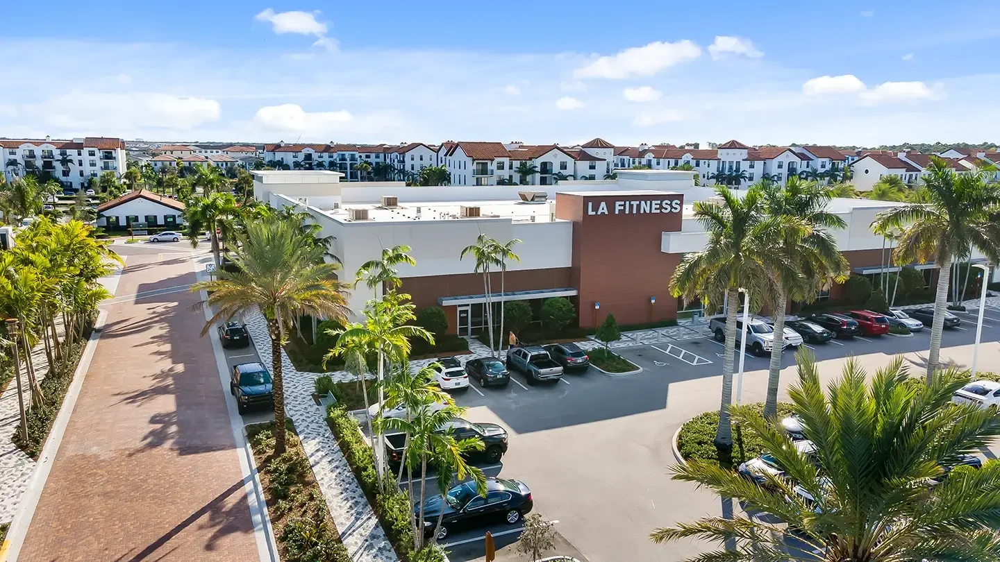 Street-level view of the LA Fitness building at Alton Town Center, featuring a brown and white exterior with clearly marked signage. The building is surrounded by palm trees, landscaping, and a small adjacent parking lot. A wide brick-paved street lined with tropical trees leads toward a residential area in the background, showcasing white multi-story buildings with red-tiled roofs under a blue sky.