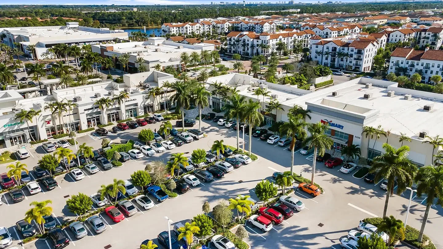 Aerial view of the PetSmart storefront at Alton Town Center, with a large parking lot filled with vehicles in the foreground. The retail plaza features white modern buildings with palm trees and greenery lining the walkways. Additional shops and signage are visible along the storefronts. In the background, multi-story residential buildings with white exteriors and red-tiled roofs can be seen, along with a lake and distant city skyline.