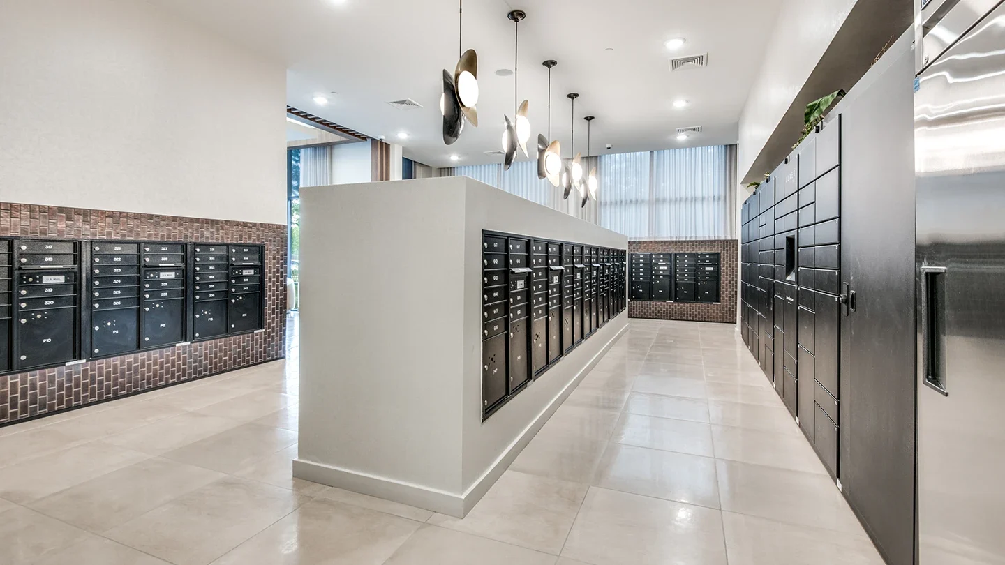 A contemporary apartment building mailroom with two rows of black mailbox units and a package locker wall. The space features neutral-colored tile flooring, brown accent brick, and decorative pendant lights hanging from the ceiling. Ample lighting and clear walkways ensure easy access for all residents.
