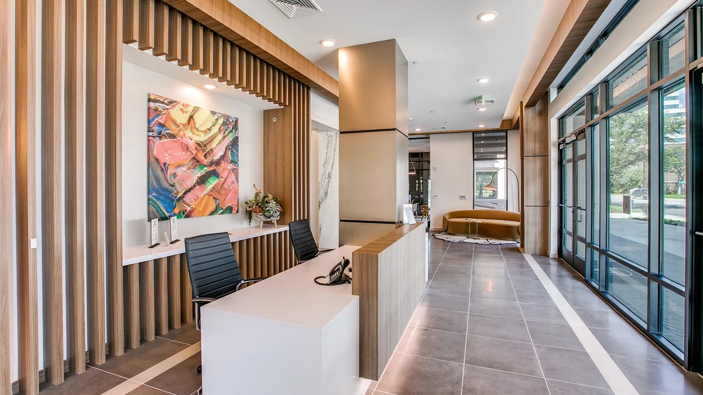 A stylish reception area with a long wood-accented front desk, two black chairs, abstract wall art, and full-height windows allowing daylight to illuminate the modern, tiled interior.