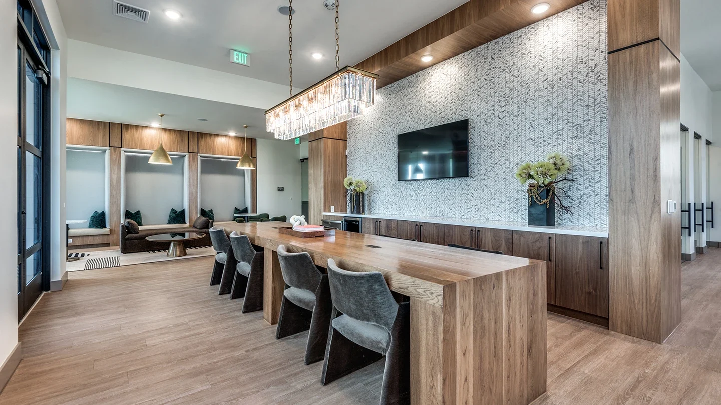 Community lounge with a large wooden communal table surrounded by velvet chairs. Features built-in wall seating, brass pendant lights, decorative wall tiles behind a mounted TV, and wood-paneled walls and cabinetry.