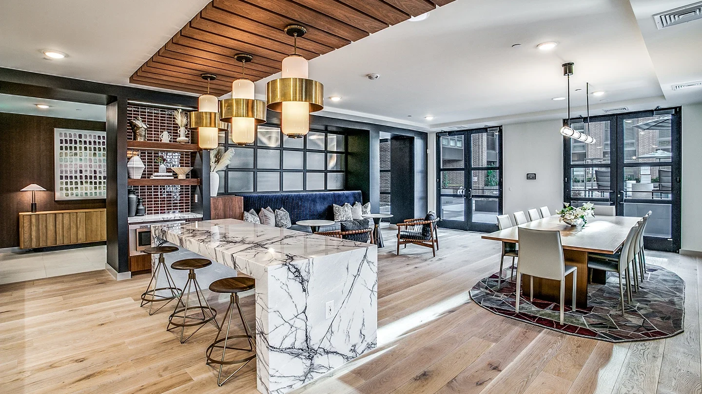 A stylish community common area with a long marble island and wooden bar stools on one side, and a large rectangular dining table with white chairs on the other. The room features warm wood flooring, pendant lighting, and a combination of built-in booth and lounge seating, creating an inviting and accessible gathering space.