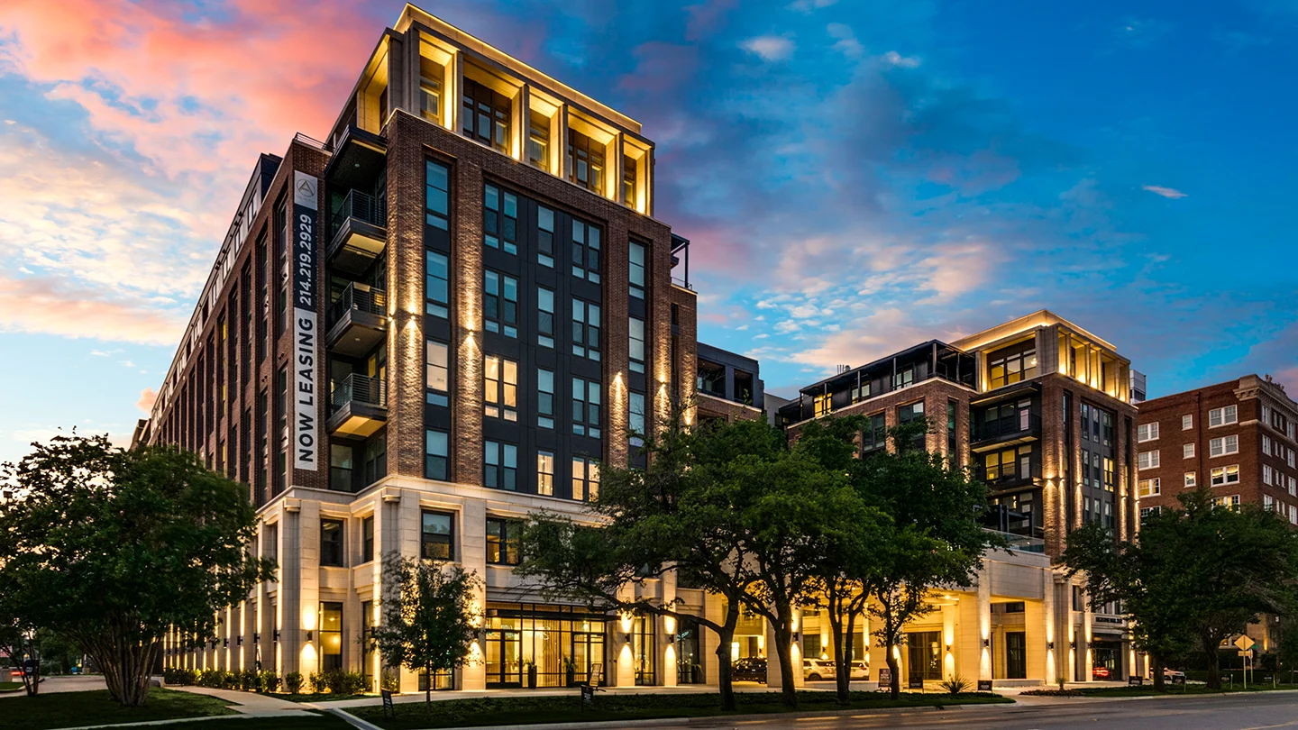 A nighttime view of a multi-story apartment building with dramatic architectural lighting highlighting the brick and stone facade. Trees and landscaping frame the front entrance, and interior lights are visible through large windows. A colorful sunset sky fades to deep blue above.