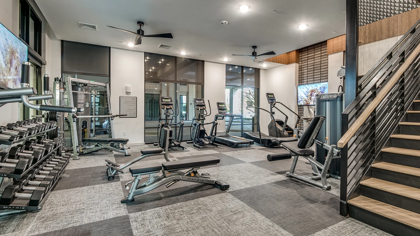 Modern fitness center with cardio and strength training equipment including treadmills, ellipticals, free weights, and benches. Room features large windows, overhead lighting, ceiling fans, and a staircase with a wood and metal railing.