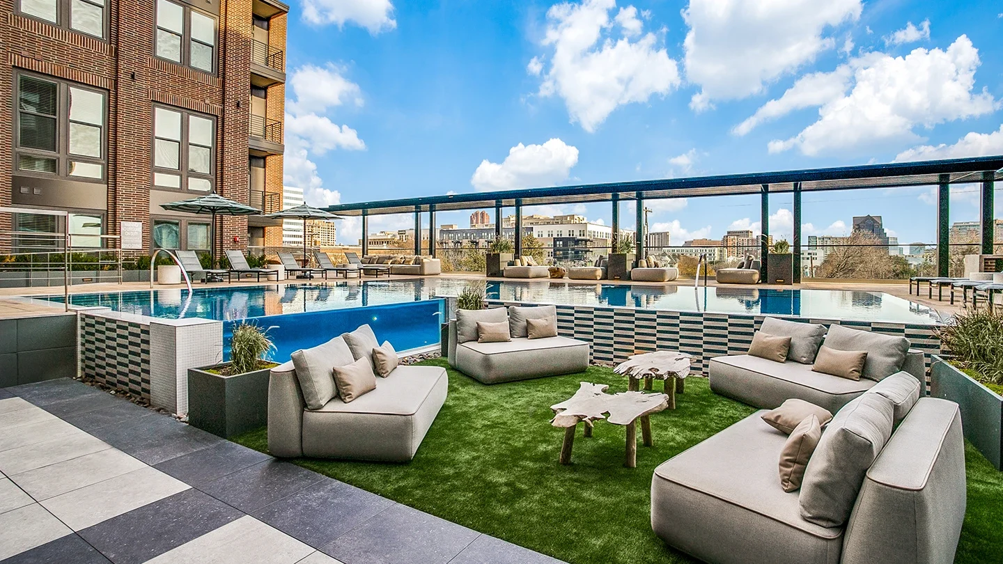 A rooftop infinity-edge pool with submerged loungers, modern outdoor seating on artificial grass, wooden accent tables, and a backdrop of city buildings under a sunny, partly cloudy sky.