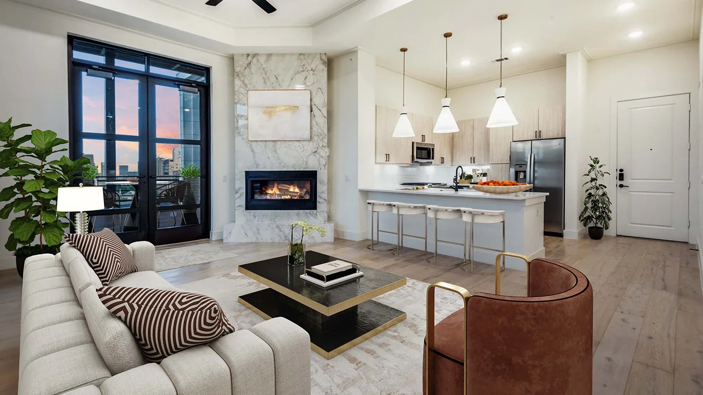 Open-concept living area with a modern kitchen featuring pendant lighting, light wood cabinetry, and a white island with bar seating. The living room includes a fireplace set in marble, a contemporary beige sofa, and large black-framed glass doors opening to a balcony with a city view at sunset.