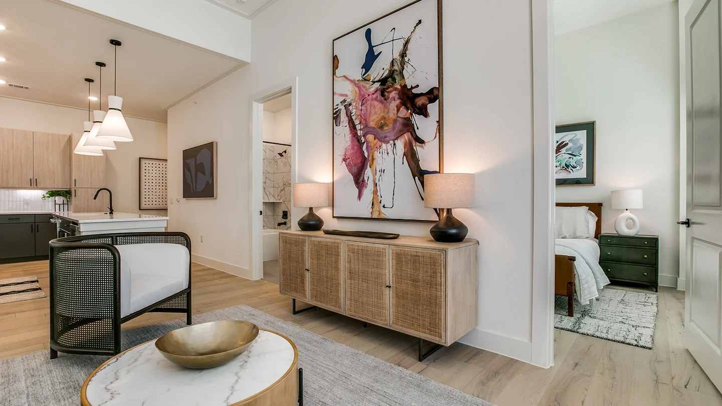 Stylish apartment interior showcasing a transition area between a modern kitchen and bedroom. Features include light wood flooring, a rattan-front console with table lamps, bold abstract artwork, and partial views of a bedroom with green nightstands and white bedding.