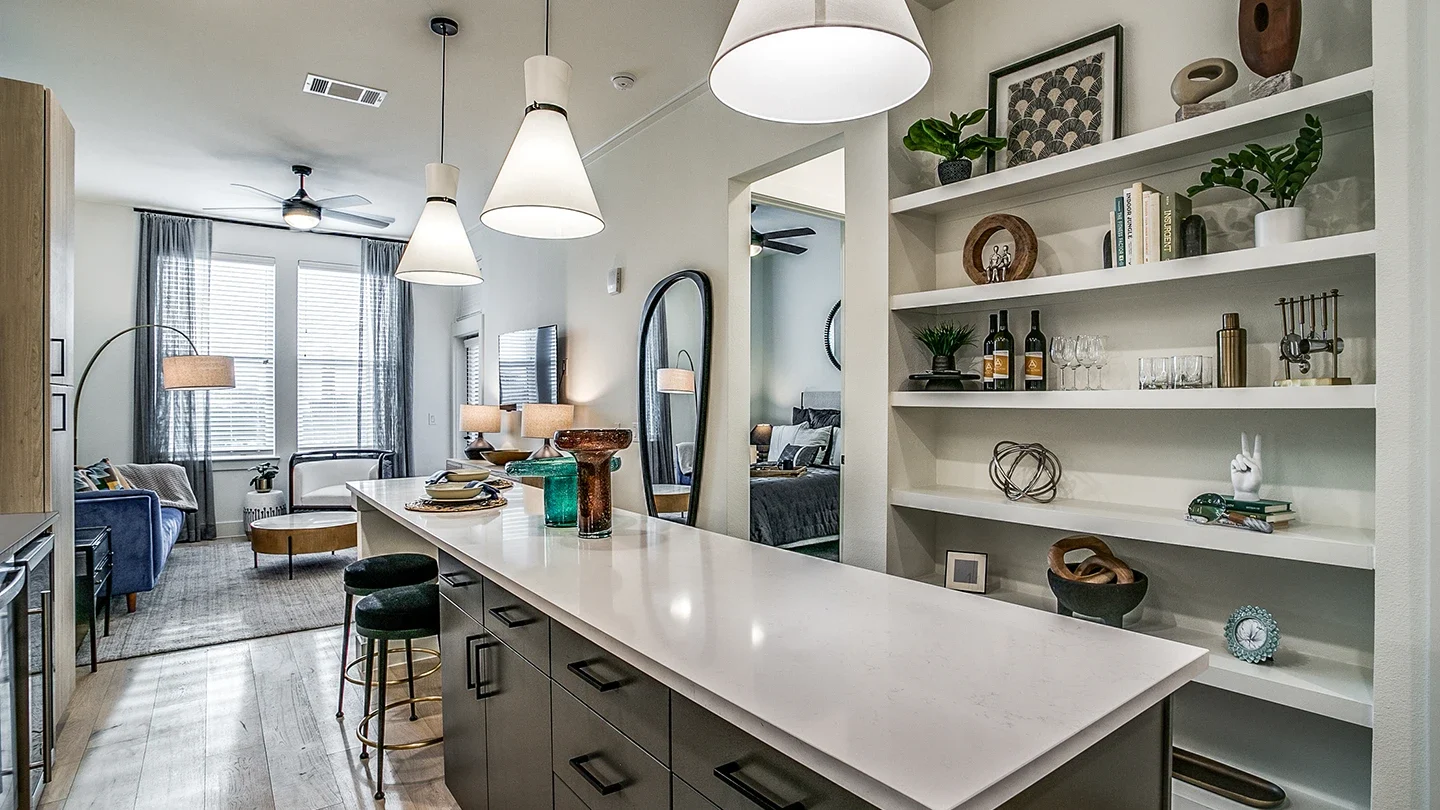Contemporary apartment kitchen with a long white island, dark lower cabinets, and decorative shelving. Open shelving displays books, plants, wine, and sculptures. In the background, a living room with blue and white furniture and a wall mirror is visible along with an adjacent bedroom.