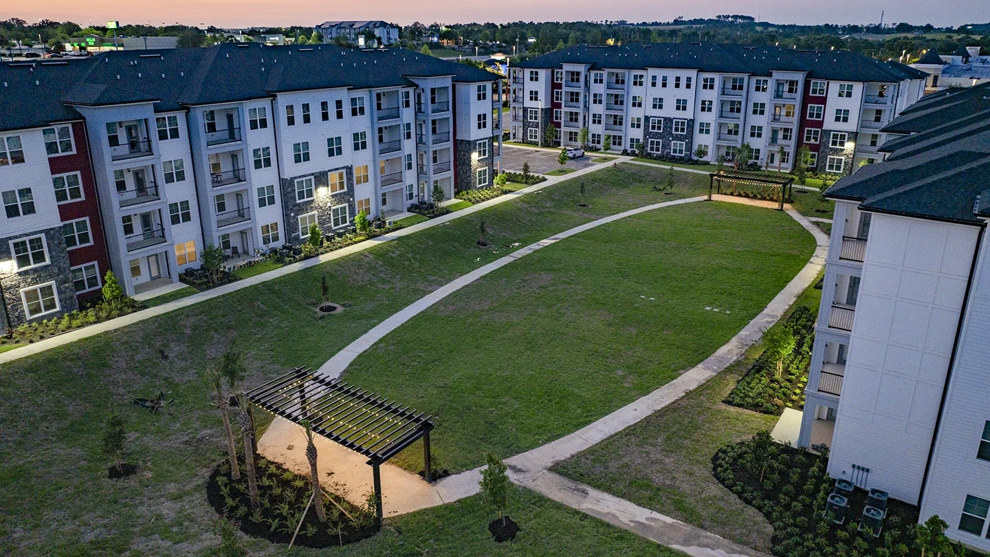 Aerial view of a large grassy courtyard surrounded by four-story apartment buildings at dusk. Curved sidewalks loop through the space, with two wooden pergolas lit from beneath. Newly planted trees line the walkways, and lights illuminate the building entrances.