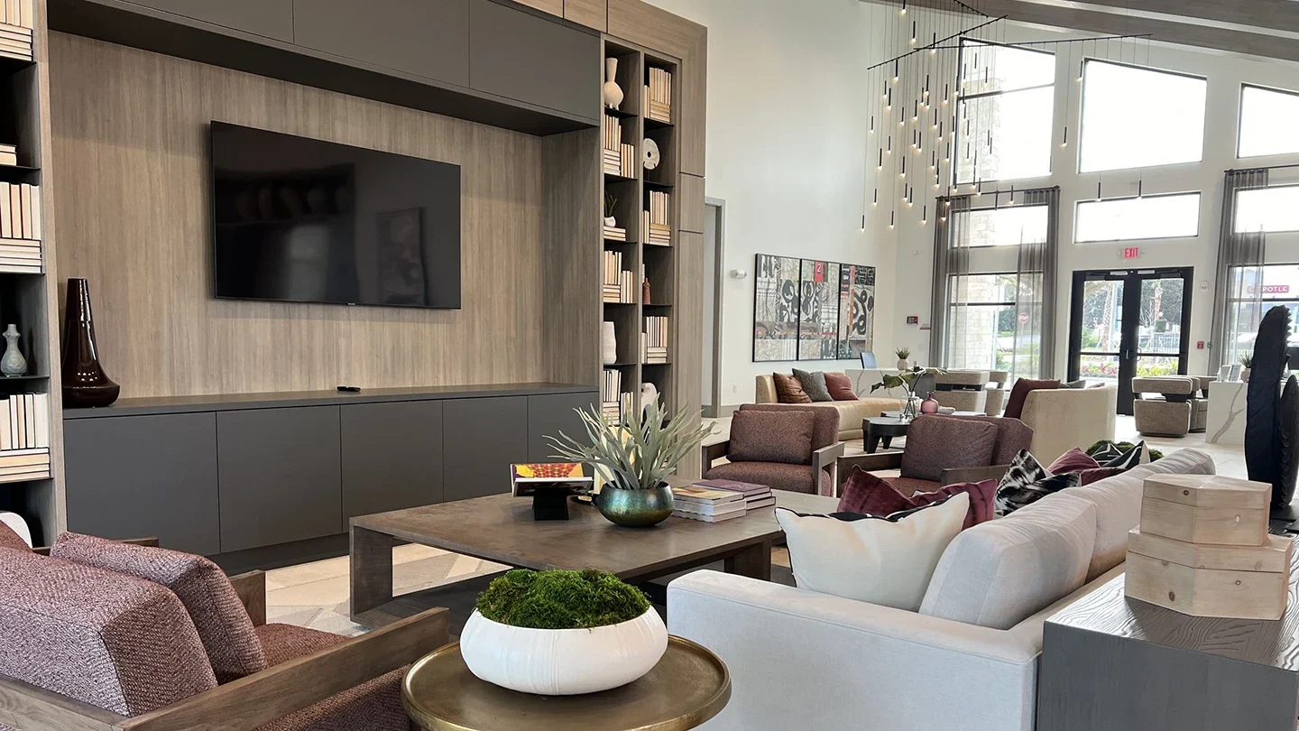Spacious and accessible community lounge with modern furniture, large windows providing natural light, and a mounted television surrounded by built-in shelving.