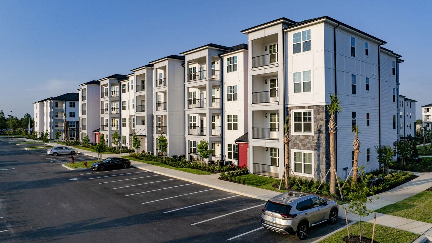 Ground-level view of a modern four-story apartment building with white panel siding, stone accents, and balconies. The building faces a paved parking lot with several cars parked along a tree-lined walkway.