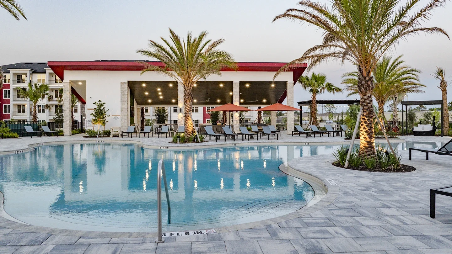 Resort-style outdoor pool area featuring a curved swimming pool surrounded by palm trees, lounge chairs, and umbrellas. A modern clubhouse with a covered patio is in the background, illuminated by ceiling lights.