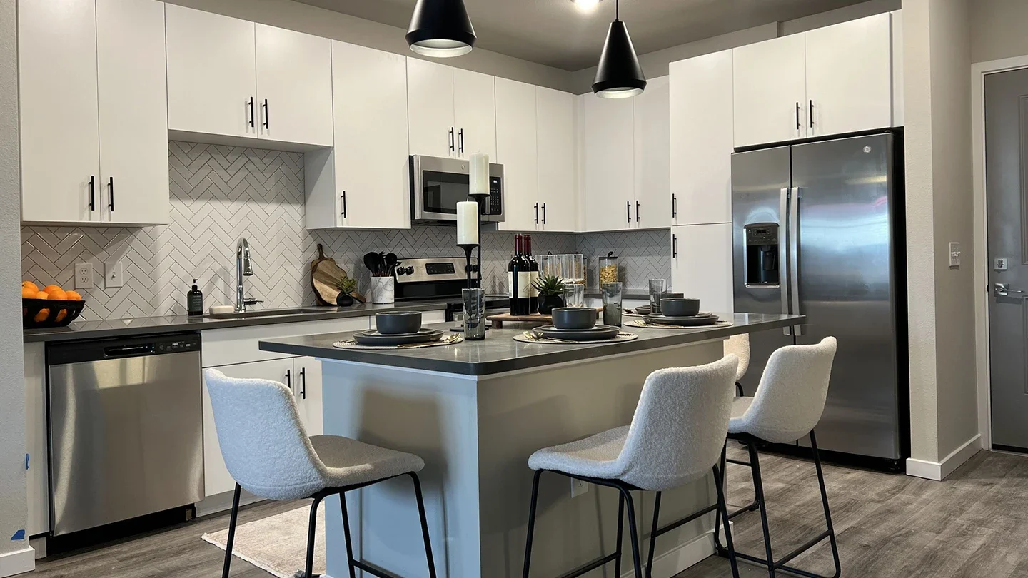 Modern kitchen with white cabinetry, stainless steel appliances, and a gray island with four upholstered bar stools. Features include a built-in microwave, dishwasher, herringbone tile backsplash, pendant lighting, and a place setting for four on the island counter.