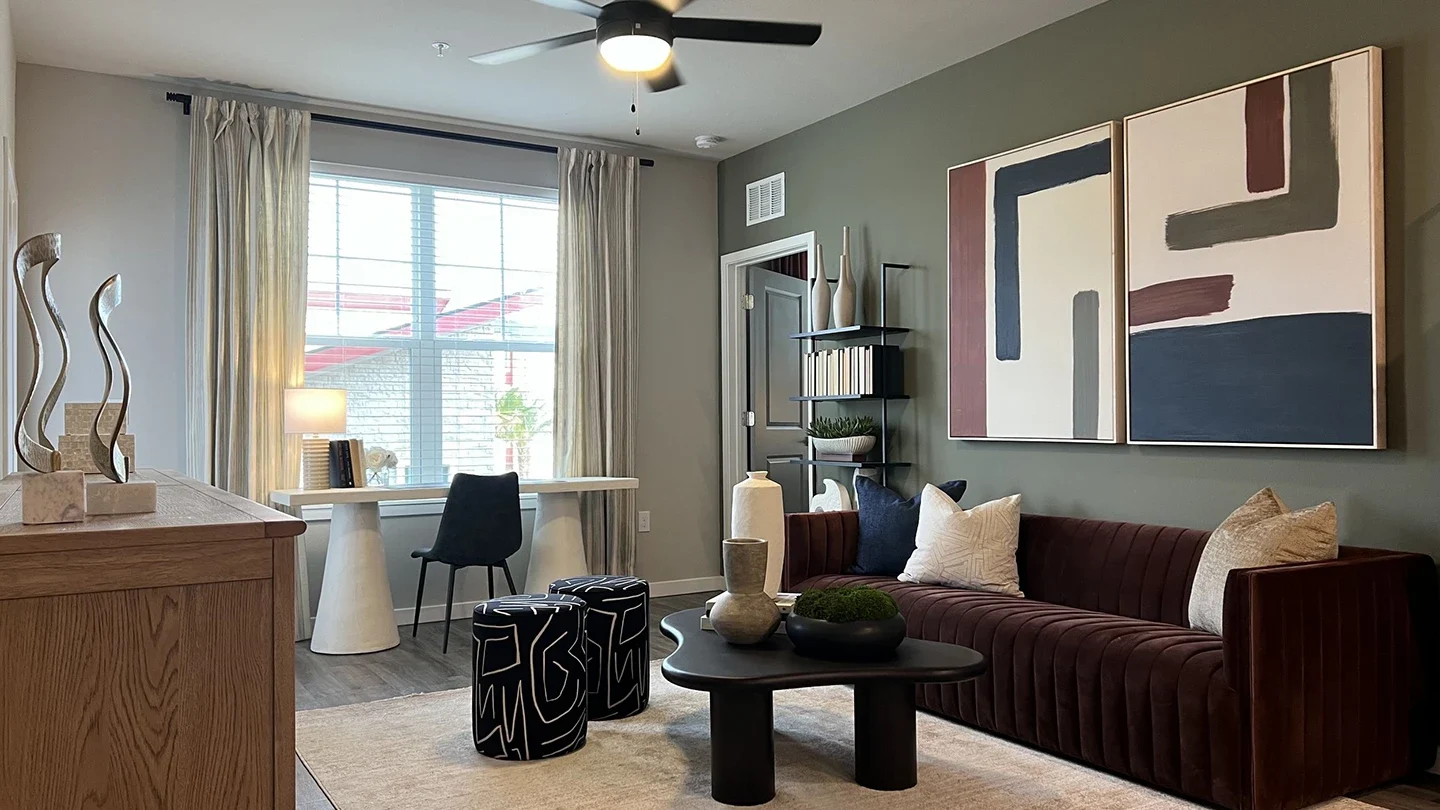 Stylish living room with a large window, dark brown velvet sofa, modern abstract artwork on the wall, and a black coffee table with decorative vases. Includes a compact desk setup with a chair near the window and two patterned ottomans in front of the couch.