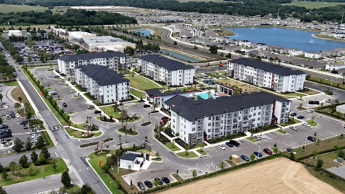 Aerial view of Ilumina Zephyrhills, a modern apartment complex featuring multiple four-story white residential buildings with dark roofs arranged around landscaped green spaces, a pool area, and parking lots. Surrounding the complex are main roads, additional parking areas, retail buildings, and residential neighborhoods with a large lake and greenery in the background.