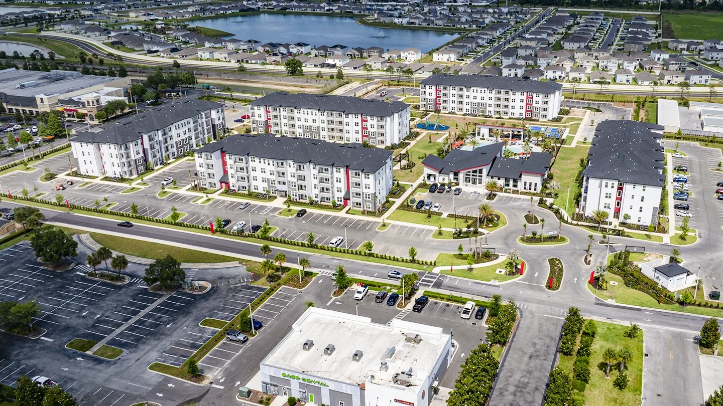 Aerial view of the Ilumina Zephyrhills apartment complex showing multiple four-story white residential buildings with black roofs, ample parking spaces, landscaped green areas, a central clubhouse, and a swimming pool. Surrounding the property are commercial buildings, roadways, and suburban neighborhoods with a lake and highway in the distance.