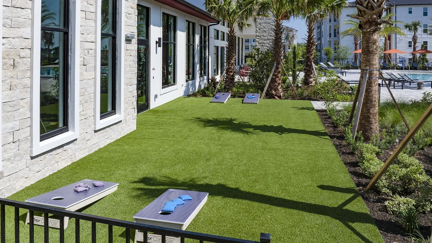 Accessible outdoor recreation area with artificial turf featuring cornhole boards, surrounded by palm trees and landscaping, adjacent to a modern building with large windows.