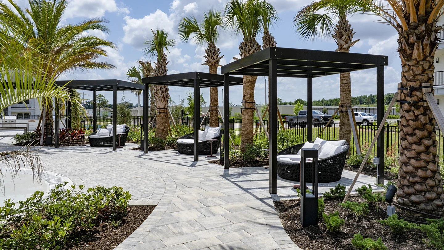Modern poolside cabanas with black metal frames and cushioned wicker lounge chairs, surrounded by palm trees and landscaping along a curved stone pathway under a bright blue sky.